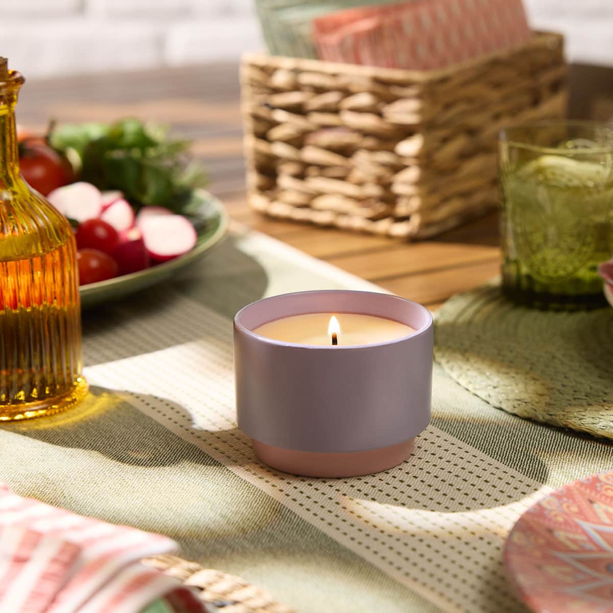 Geranium Citronella™ Two-Tone Jar Candle on a woven table mat on a wooden table with bowls fruit and a wicker box with papers in the background