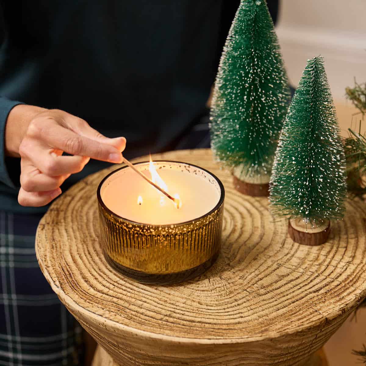Whiskey & Oak 3-Wick Ripple Jar Candle being lit with a long match stick by a lady hand on a wood surface with decorative glittered tips Christmas Trees.