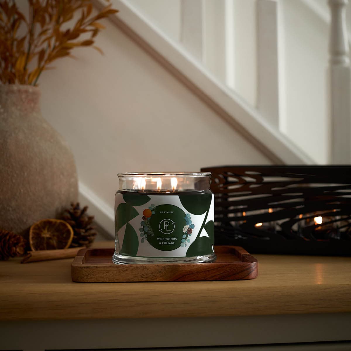 Wild Woods & Foliage 3-Wick Jar Candle lit on a wooden surface with white stair bannisters in the background.