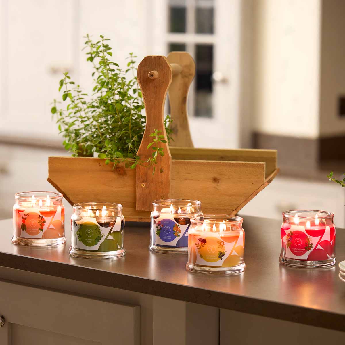Assorted colorful 3-wick Jar Candles on a wooden surface with a wooden plant pot with a plant in the background.